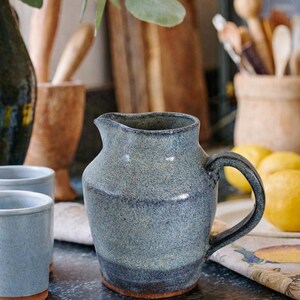 May include: A handmade ceramic pitcher with a dark blue and gray speckled glaze. The pitcher has a curved handle and a spout for pouring. The base is a darker shade of blue. Other ceramic items and lemons are in the background.