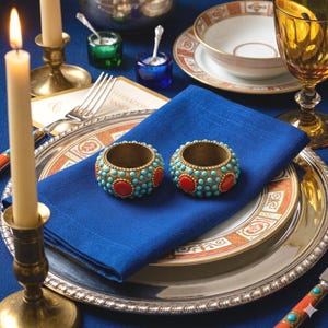 May include: Two circular napkin rings featuring a metallic base adorned with small, round turquoise beads and larger, smooth oval red stones. The rings are positioned on a folded deep blue cloth napkin resting on a silver-rimmed dinner plate.