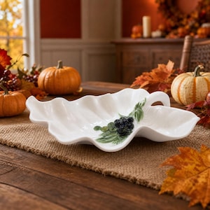 May include: A white ceramic serving dish shaped like a large, wavy leaf. The dish features a raised, three-dimensional decorative accent of dark purple grapes and green leaves on one side, with a curved handle integrated into the leaf design.