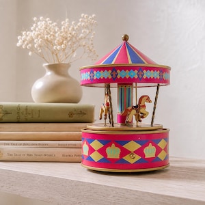 May include: A decorative carousel music box featuring a pink and blue striped conical roof, a central pillar, and miniature horses suspended by twisted metal poles. The base is decorated with a pink, blue, and yellow diamond pattern and gold crowns.