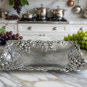 May include: A rectangular silver-toned metal serving tray featuring a raised, detailed relief pattern of grape clusters and vine leaves along the edges. The tray has a smooth, polished center surface and an ornate, textured border design.