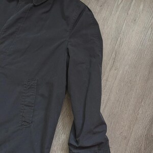 May include: A solid black long-sleeve coat featuring a classic pointed collar, a button-down front placket, and a single vertical side pocket. The garment is constructed with visible seam detailing along the shoulder and sleeve areas.