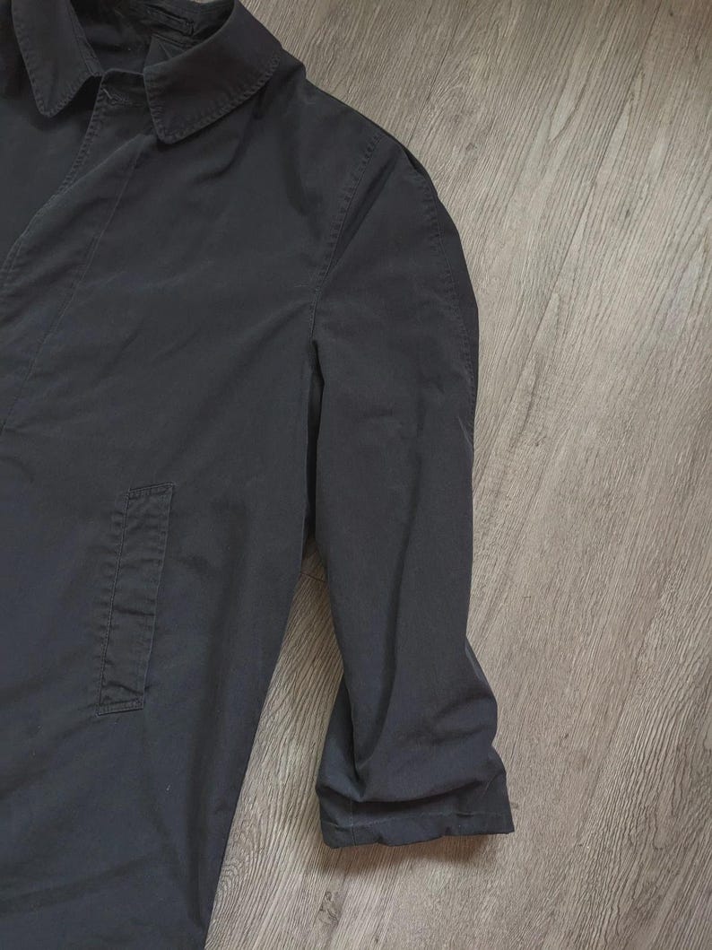 May include: A solid black long-sleeve coat featuring a classic pointed collar, a button-down front placket, and a single vertical side pocket. The garment is constructed with visible seam detailing along the shoulder and sleeve areas.
