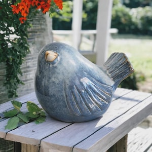 May include: A ceramic bird sculpture in shades of blue and gray sits on a wooden surface. The bird has a rounded body, textured wings, and a small, light-colored beak. The sculpture is a decorative item.