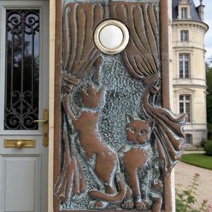 May include: Bronze doorbell with a relief of two cats and draped curtains. The doorbell button is white and round. The cats are in a playful pose, one reaching up. The doorbell is mounted on a stone wall.