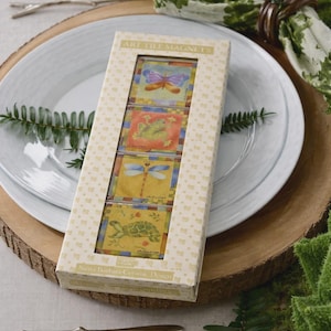 May include: A decorative box of art tile magnets featuring colorful insect and reptile designs. The box includes a butterfly, frog, dragonfly, and turtle. The box is on a wooden charger with a white plate and a green leaf napkin.