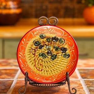 May include: A decorative ceramic grater plate with a vibrant orange rim and a yellow center. The plate features a textured grating surface and is supported by a wrought iron stand. The design includes floral patterns and dark green accents.