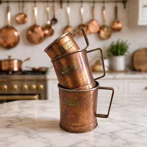 May include: Three antique copper measuring cups stacked on a marble countertop. Each cup has a rectangular label that reads "GRAIN". The cups have brass handles and are set against a backdrop of copper cookware.