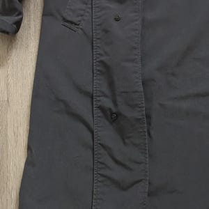 May include: A dark charcoal grey trench coat featuring a long, structured silhouette with a vertical placket covering the front closure. The garment includes visible black snap buttons along the placket and a long sleeve with a cuff detail.