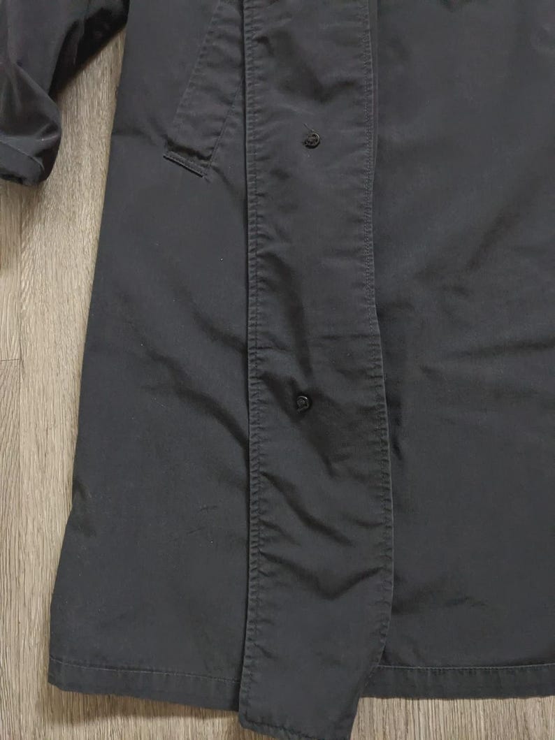May include: A dark charcoal grey trench coat featuring a long, structured silhouette with a vertical placket covering the front closure. The garment includes visible black snap buttons along the placket and a long sleeve with a cuff detail.