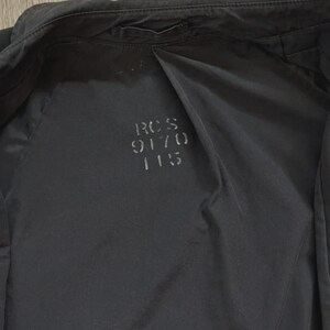May include: A black jacket with a smooth, matte finish. The interior lining features the printed text RCS 9170 115 in a dark grey font. The garment includes a collar, long sleeves, and a central seam running down the back panel of the interior.