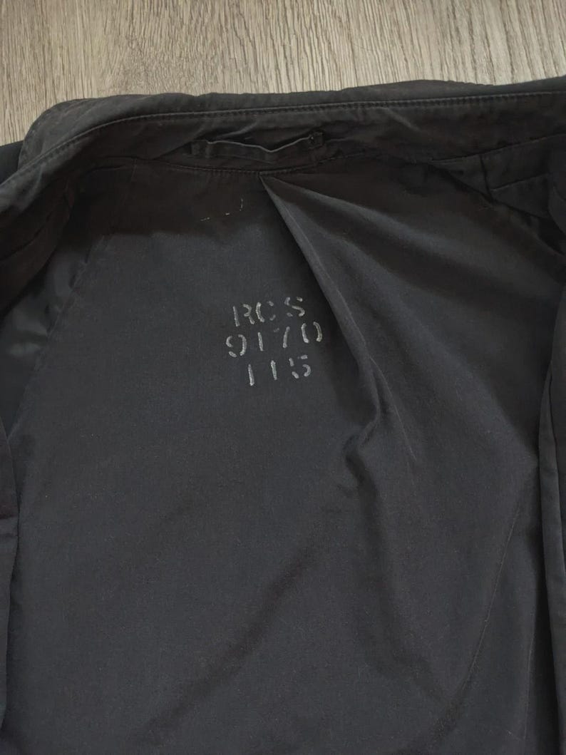 May include: A black jacket with a smooth, matte finish. The interior lining features the printed text RCS 9170 115 in a dark grey font. The garment includes a collar, long sleeves, and a central seam running down the back panel of the interior.