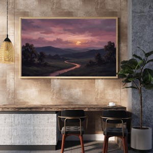 Könnte beinhalten: Ein gerahmtes Landschaftsgemälde zeigt einen Sonnenuntergang über einem Tal mit einem sich schlängelnden Fluss. Der Himmel ist eine Mischung aus rosa und lila Farbtönen, wobei die Sonne hinter den fernen Bergen untergeht. Das Gemälde wird über einem Holztisch mit zwei Stühlen ausgestellt.