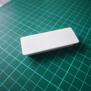 May include: A small, rectangular, white plastic object with a slightly textured surface. The object is resting on a green cutting mat with a grid pattern.