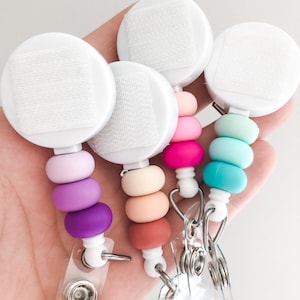 May include: Four badge reels with white circular tops and colorful silicone beads in shades of purple, pink, peach, and teal. Each reel has a clear plastic strap and a metal clip for attaching to clothing.