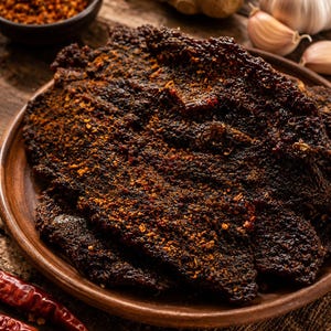 May include: A dark brown, seasoned beef jerky on a round brown ceramic plate. The meat has a textured, dried surface coated with reddish-orange spice flakes. Nearby are whole garlic cloves, a piece of ginger, a small bowl of chili flakes, and red chilies.