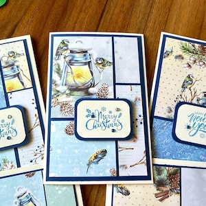 Könnte beinhalten: Eine Sammlung rechteckiger Grußkarten mit winterlichen Illustrationen von Laternen, Vögeln, Tannenzapfen und Federn. Die Karten sind mit dunkelblauen Rändern versehen und tragen kleine rechteckige Etiketten mit dem Text Merry Christmas oder Happy New Year.