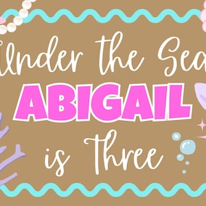 May include: A rectangular sign with a brown background and a light blue wavy border. The text reads Under the Sea ABIGAIL is Three in white and pink. Decorations include a pink starfish, a purple seashell, a mermaid tail, and white pearls.