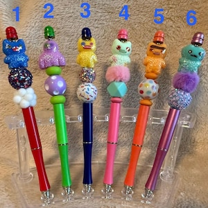 Könnte beinhalten: Sechs Stifte in einer Reihe, jeder mit einer einzigartigen Monsterfigur und dekorativen Perlen verziert. Die Stifte haben leuchtende Farben wie Rot, Grün, Blau, Pink, Orange und Lila sowie verschiedene Texturen wie Glitzer, Pompons und Punkte.