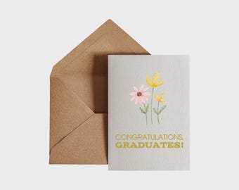 May include: A graduation card with a watercolour floral design, featuring pink and yellow flowers with green stems. The card reads "CONGRATULATIONS, GRADUATES!" in gold lettering, with a brown paper envelope.