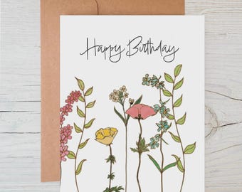 May include: A white birthday card with the words "Happy Birthday" in cursive. The card has a floral illustration with pink, yellow, and blue flowers. A brown envelope is behind the card.