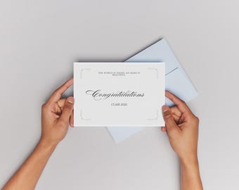 May include: A white graduation card with the word "Congratulations" in elegant script, along with the text "CLASS 2026" and the quote "THE WORLD IS YOURS, GO MAKE IT BEAUTIFUL." The card is held over a light blue envelope.