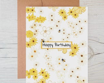 May include: A birthday card with a floral design featuring yellow and white flowers and bees. The words "Happy Birthday" are in a gold rectangle. The card is paired with a brown envelope.