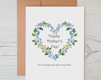 May include: A white Mother's Day card featuring a heart-shaped wreath of blue flowers and green leaves. The text "Happy Mother's Day!" is inside the heart, with the phrase "I'm so lucky that you're my mom." below. A brown envelope is behind the card.
