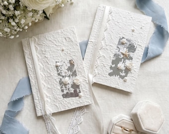 May include: Two white textured paper notebooks decorated with silver foil accents, small white fabric flowers, pearls, and a starfish. A white lace trim and ribbon are attached to the covers. A beige velvet hexagonal ring box holds two gold rings.