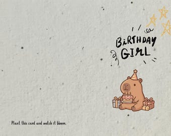 May include: Birthday card featuring a cartoon capybara with a cake and candles. The card reads "Birthday Girl" and "Plant this card and watch it bloom." The background is a textured, off-white colour.