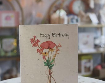 May include: A birthday card with the words "Happy Birthday" in black script. The card features a watercolour illustration of a bouquet of flowers, including a red poppy, yellow daisies, and other wildflowers. The card is on a white surface.