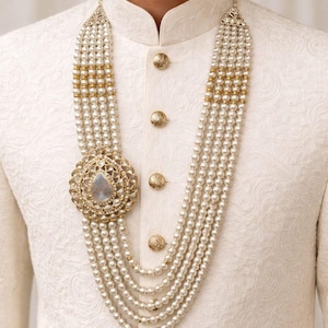 May include: A multi-strand pearl necklace with a large, ornate pendant. The pendant features a central teardrop-shaped stone surrounded by a gold-toned filigree design. The necklace is worn over a white, textured garment with gold buttons.