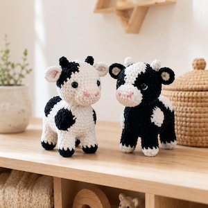Könnte beinhalten: Zwei handgefertigte Häkelfiguren von Kühen. Eine Kuh ist weiß mit schwarzen Flecken, die andere schwarz mit weißen Flecken. Beide haben rosa Nasen und kleine schwarze Augen. Sie werden auf einem Holzregal ausgestellt und zeigen eine charmante, handwerkliche Ästhetik.