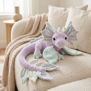 以下が含まれることがあります： ラベンダー、ミントグリーン、クリーム色の手編みドラゴンぬいぐるみ。 大きな翼、長い尻尾、フレンドリーな表情が特徴です。 柔らかいおもちゃはベージュのソファの上に置かれています。