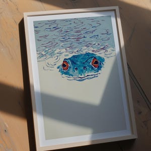 以下が含まれることがあります： 水から現れる赤い目の青いカエルを描いた額入りのアートプリント。アートワークは、木製のフレームで、明るい青と白の配色を使用しています。カエルの顔が焦点です。