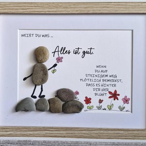 Puede incluir: Obra de arte enmarcada con una figura de arte de guijarros sosteniendo una flor. El texto "Alles ist gut" está escrito encima de la figura. La obra de arte está en un marco de madera clara.