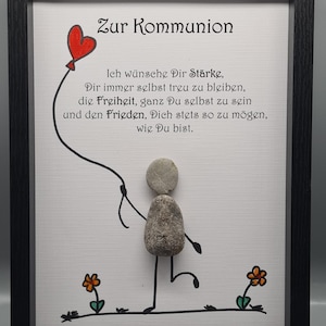 Puede incluir: Obra de arte enmarcada con el texto alemán "Zur Kommunion" y un mensaje. La obra presenta una figura de palo hecha de piedras que sostiene un globo de corazón rojo. El marco es negro.