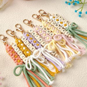 Peut inclure: Une collection de six porte-clés personnalisés avec des motifs de fleurs au crochet dans des tons pastel, des perles en bois avec des lettres formant les prénoms Hannah, Robert, Emma, Crist, Ally et Katil, des mousquetons dorés et des pompons en corde.