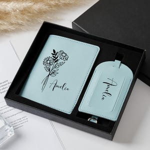 Puede incluir: Un juego de funda para pasaporte y etiqueta de equipaje en color azul claro, presentado en una caja de regalo negra. La funda incluye un gráfico floral negro y el nombre Amelia en cursiva. La etiqueta también muestra el nombre Amelia.