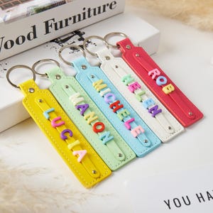 May include: Four rectangular keychains in yellow, light green, light blue, and red, each featuring a silver metal ring and a central strap. The keychains display the names LUCIA, MANON, MICHELE, HELEN, and ROB in colourful, raised block letters.