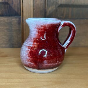 May include: A small, ceramic pitcher with a rounded body and a handle. The pitcher is primarily a deep red color, with a white rim and a white base. The pitcher is sitting on a light brown wooden surface.