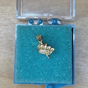 May include: A gold-colored pin with a crown and a red gem, featuring the word "WEIRD". The pin is displayed in a blue box with a clear lid, set against a wooden surface. The pin is likely a piece of jewelry or a collectible item.