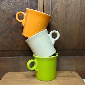 May include: Three ceramic mugs stacked on top of each other. The top mug is orange, the middle mug is white, and the bottom mug is green. Each mug has a rounded handle. The mugs are on a wooden surface with a wooden background.