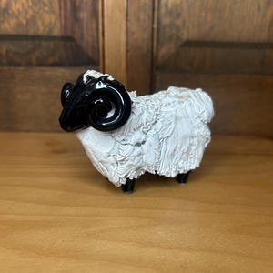 May include: A ceramic ram figurine with a black head and horns, and a white, textured body. The ram has four black legs and is set against a wooden background.