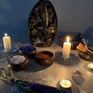 May include: A low-lit scene with an incense waterfall fountain depicting a mountain scene. Small wooden bowls contain crystals, salts, and other items. Lit candles and herb bundles are also present, creating a spiritual or meditation theme.