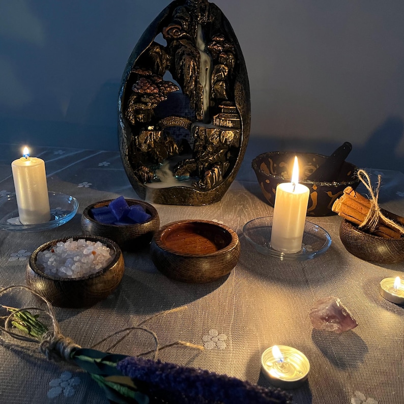 May include: A low-lit scene with an incense waterfall fountain depicting a mountain scene. Small wooden bowls contain crystals, salts, and other items. Lit candles and herb bundles are also present, creating a spiritual or meditation theme.