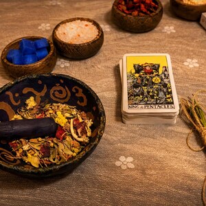 K&ouml;nnte beinhalten: Ein Stillleben mit M&ouml;rser und St&ouml;&szlig;el mit getrockneten Kr&auml;utern, kleinen Holzschalen mit blauen W&uuml;rfeln und Salz sowie einem Tarotkartenspiel mit der Aufschrift "King of Pentacles". Ein Kr&auml;uterb&uuml;ndel und eine Schachtel sind ebenfalls vorhanden.