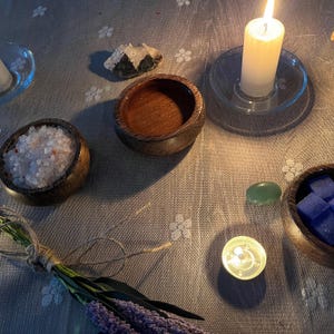 May include: A tranquil arrangement with a lit white candle on a glass stand, wooden bowls containing salt and blue cubes, and a small green stone. A bundle of lavender and a lit tea light contribute to the peaceful setting. The items are set on a floral patterned tablecloth.