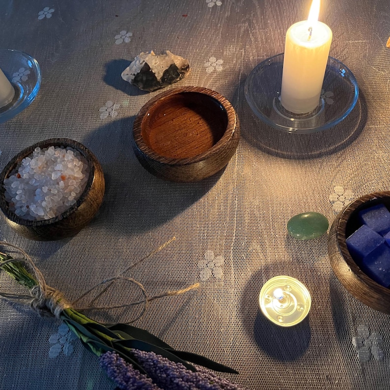 May include: A tranquil arrangement with a lit white candle on a glass stand, wooden bowls containing salt and blue cubes, and a small green stone. A bundle of lavender and a lit tea light contribute to the peaceful setting. The items are set on a floral patterned tablecloth.