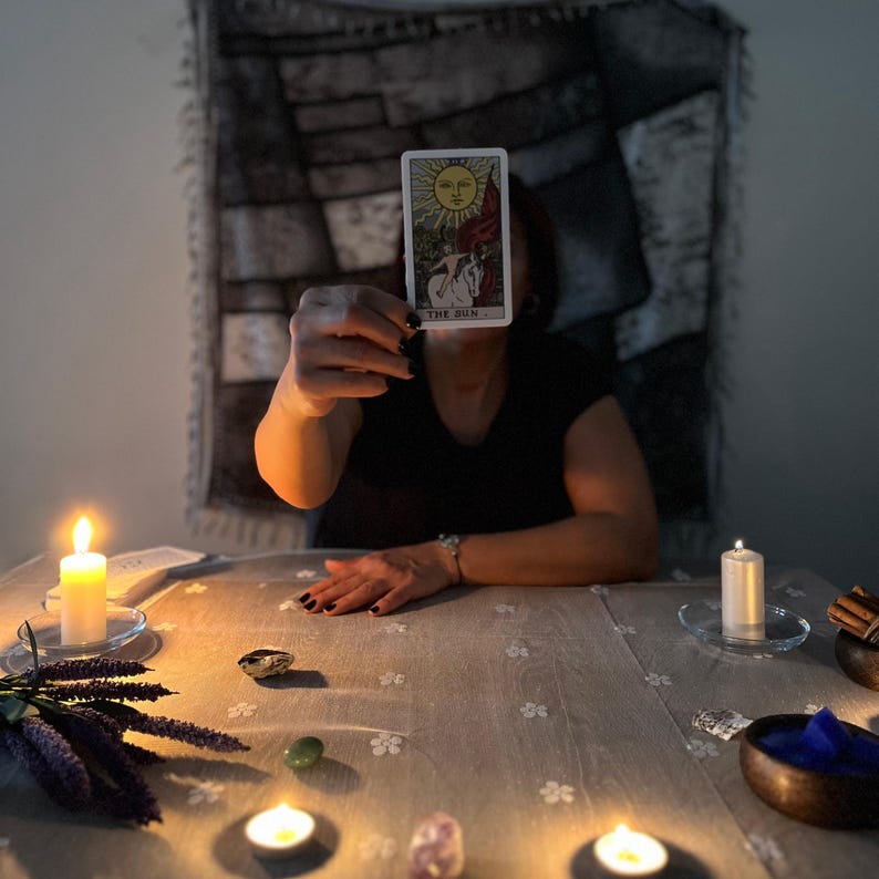 May include: A tarot card reading scene with a person holding up a card labelled "The Sun." The card features a yellow sun with a face, a white horse, and a red flag. Lit candles and crystals are on the table, creating a mystical atmosphere.